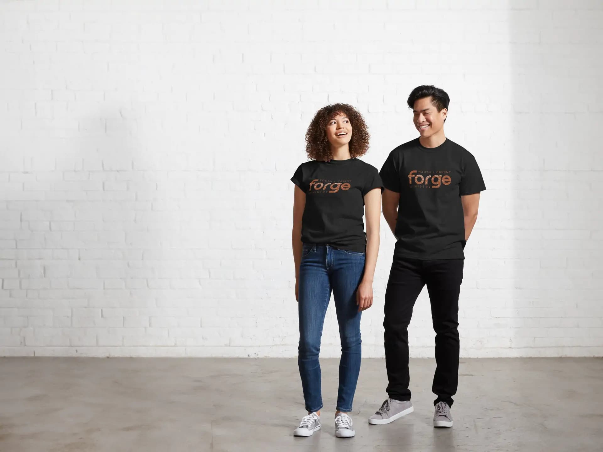 A Black woman and Asian man standing next to each other, smiling, while both wearing blue t-shirts featuring the logo for Forge – Youth and Parent Ministry, designed by Andrilla.